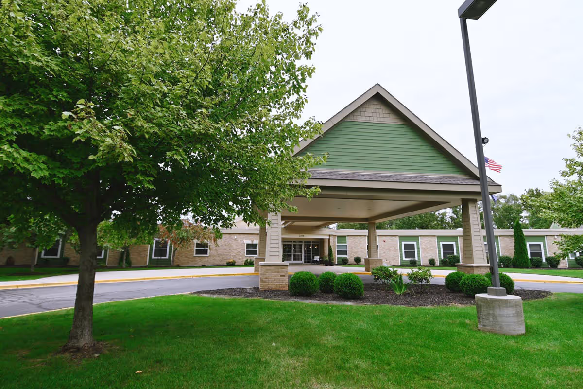 Exterior view of The Cortland Wyoming senior living facility showing the main entrance with a covered driveway, surrounded by green grass, bushes, and trees under an overcast sky.