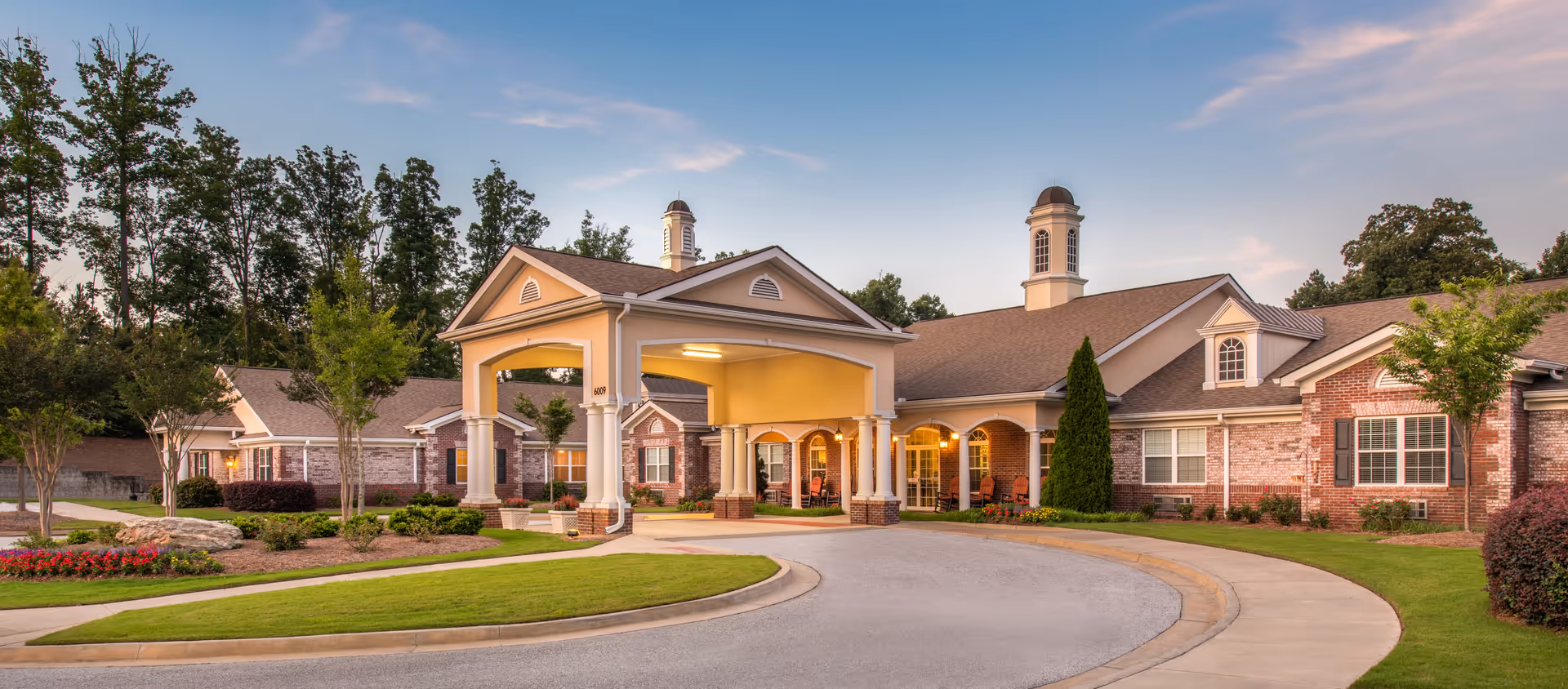 Exterior view of Benton House of Staley Hills, a single-story senior living facility with a covered entrance, brick and siding facade, surrounded by landscaped greenery and trees under a clear sky.