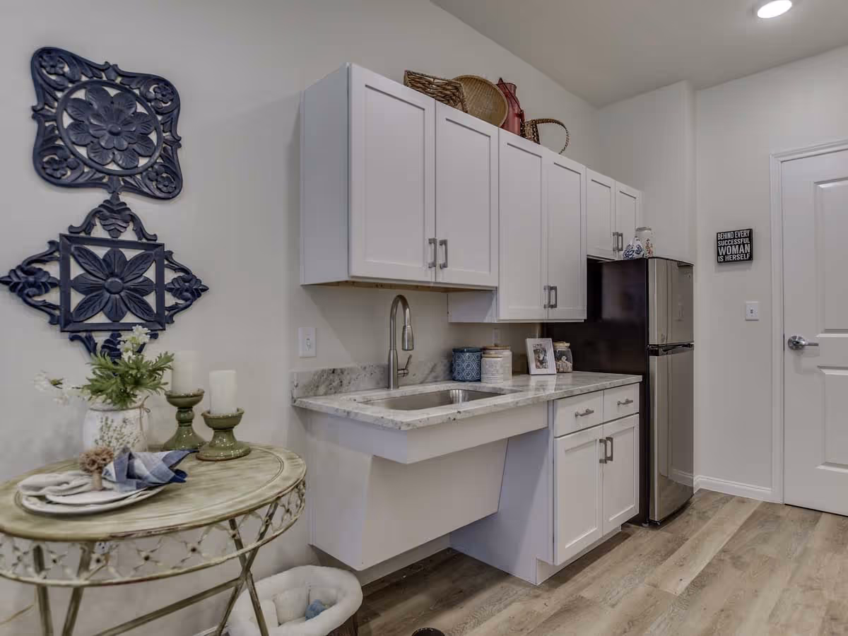 Small kitchenette with white cabinets, a marble countertop and sink, a stainless steel mini fridge, decorative wall art, and a round side table with candles and flowers.