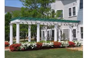 Outdoor view of a white pergola structure with green lattice roofing in front of a multi-story white building. The pergola is surrounded by neatly maintained flower beds with red and white flowers and green grass.