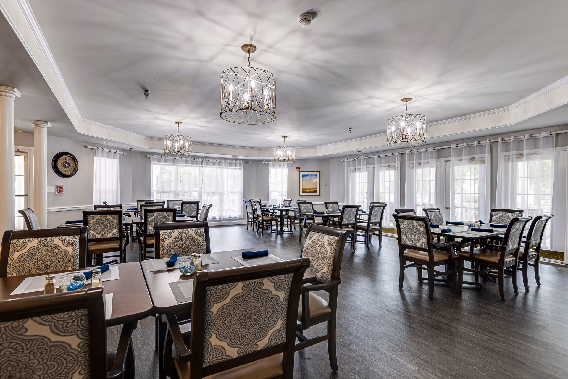 A spacious dining room with multiple tables and chairs arranged neatly. The chairs have patterned upholstery, and the tables are set with placemats, napkins, and small decorative items. Large windows with sheer white curtains allow natural light to fill the room. Elegant chandeliers hang from the ceiling, casting soft light and shadows. The room has a clean, modern design with light-colored walls and dark wood flooring.