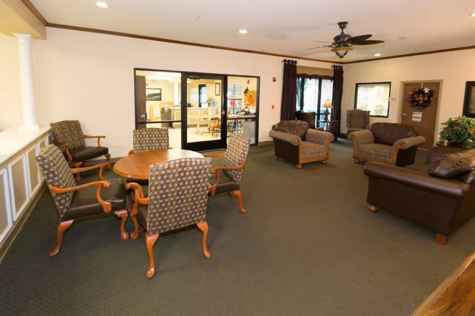 A spacious living room area with a round wooden table surrounded by four patterned chairs with wooden arms. There are additional cushioned armchairs and a leather sofa arranged around the room. The room has beige walls, carpeted floor, a ceiling fan with light, and large windows with dark curtains. A glass door leads to another room in the background.