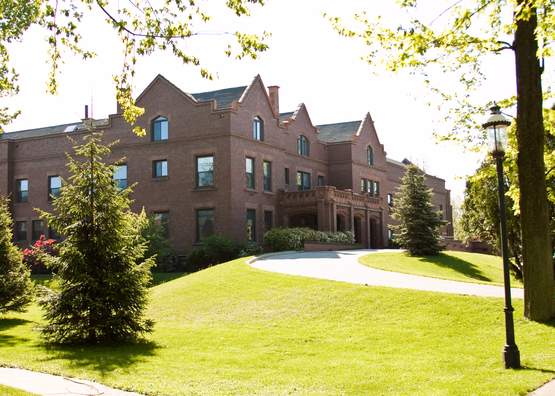 A large brick building with multiple windows and a peaked roof, surrounded by green grass, trees, and a curved driveway leading to the entrance. A lamppost is visible on the right side of the image.