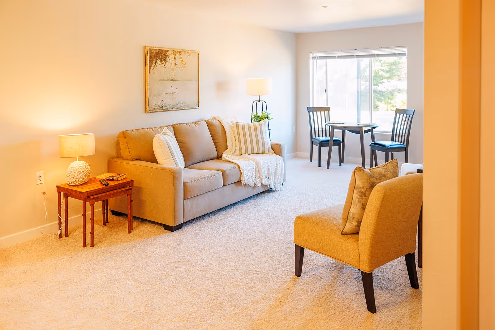 A bright living room with beige carpet and walls, featuring a beige sofa with white and striped pillows and a throw blanket. There is a wooden side table with a lamp and books next to the sofa. A yellow armchair with a patterned pillow is in the foreground. In the background, a small dining table with two chairs is positioned near a large window letting in natural light. A floor lamp and a framed picture hang on the wall above the sofa.