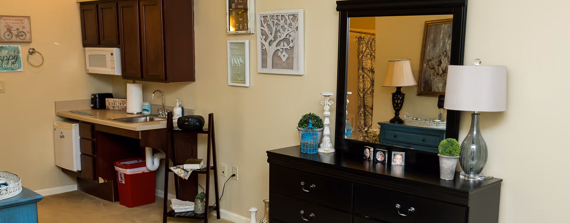 Interior view of a senior living facility room at Bickford of Oswego showing a small kitchenette with a sink, microwave, and mini fridge on the left. On the right, there is a black dresser with a large mirror, a table lamp, small decorative plants, and framed photos. The wall has several decorative art pieces, and a small shelving unit holds a radio and books.