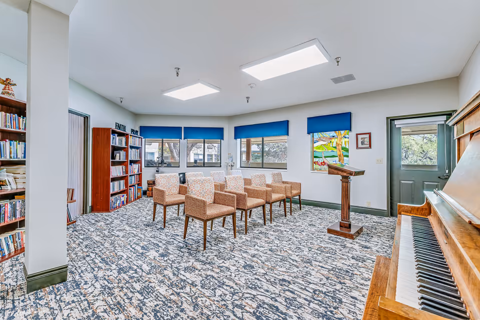 A bright room with patterned carpet and several upholstered chairs arranged in rows facing a wooden podium. There is a piano on the right side and bookshelves filled with books on the left. The room has multiple windows with blue valances and a colorful stained glass window. The walls are white with green trim, and there is a door on the right side.