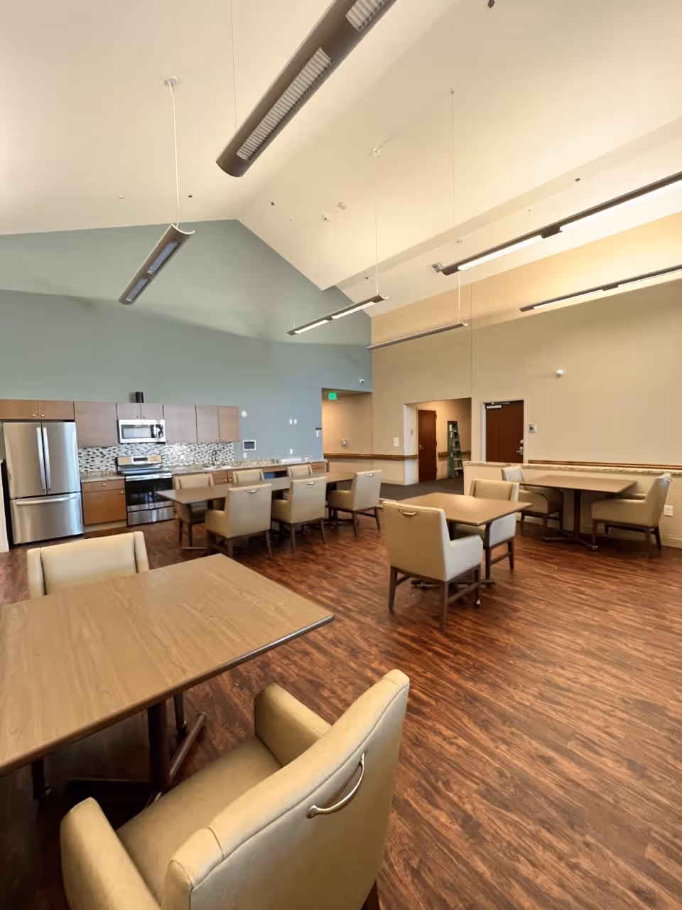 A spacious dining area with several tables and cushioned chairs arranged on a wood floor. The room features a modern kitchen along one wall with stainless steel appliances including a refrigerator, oven, and microwave. The walls are painted in light neutral tones with a high ceiling and hanging light fixtures.