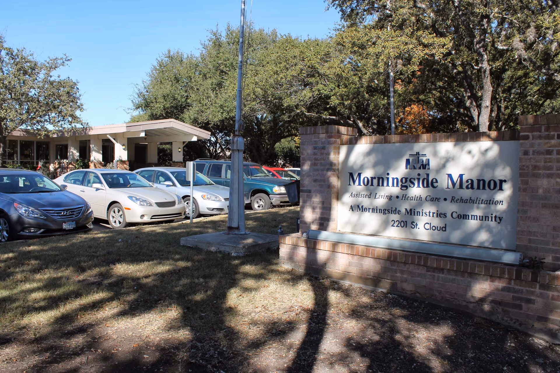 Exterior view of Morningside Manor assisted living facility showing a brick sign with the facility name and services offered, several parked cars, and trees in the background.