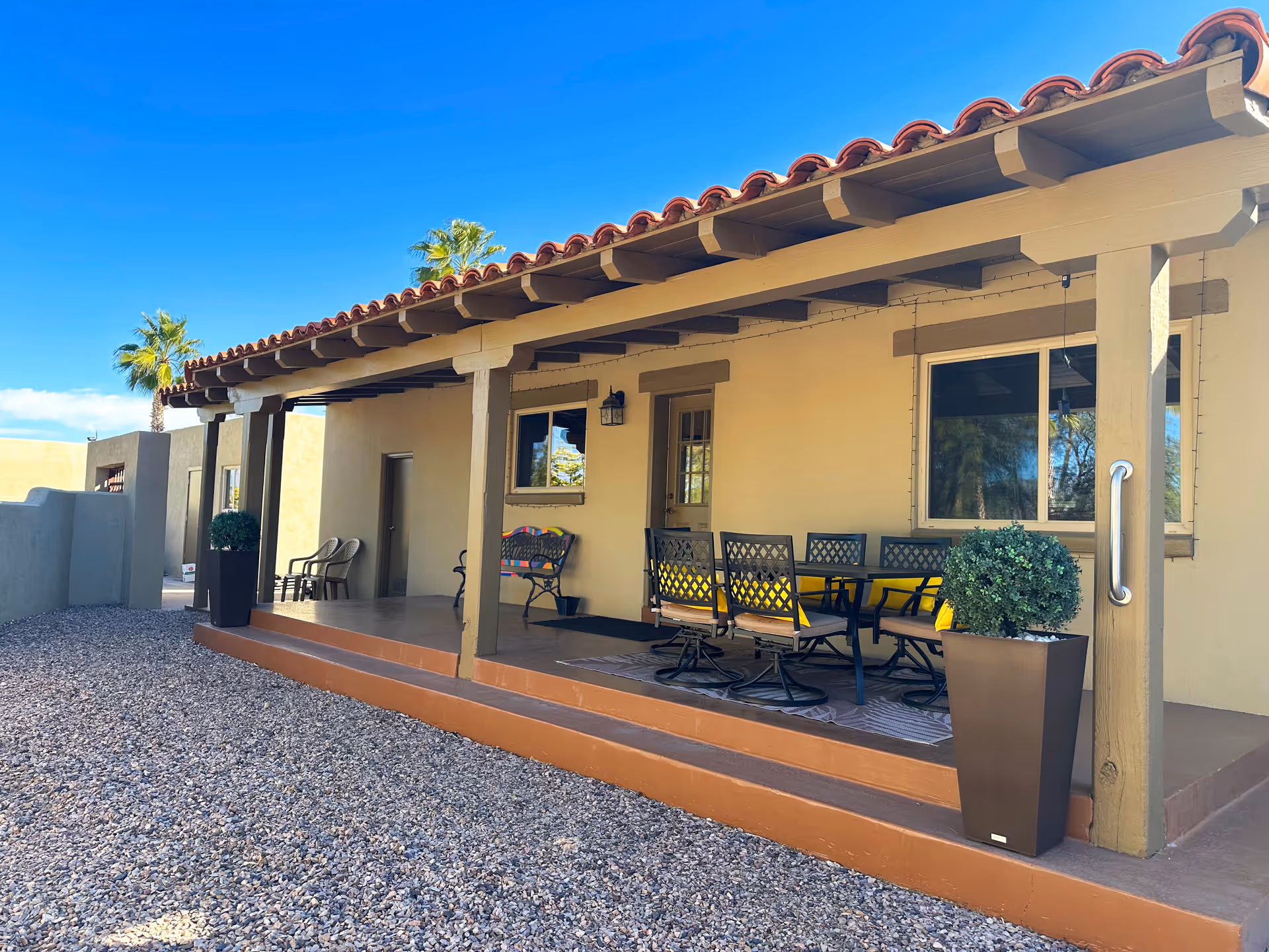 Covered outdoor patio with seating and dining table in front of a single-story stucco building with a clay tile roof.