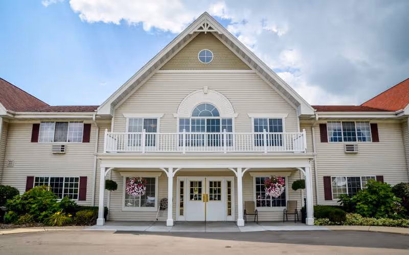 Front exterior view of a large, two-story building with beige siding, white trim, and a peaked roof. The building features a balcony with white railings above the main entrance, which has double doors and is flanked by hanging flower baskets and benches. The sky is partly cloudy.