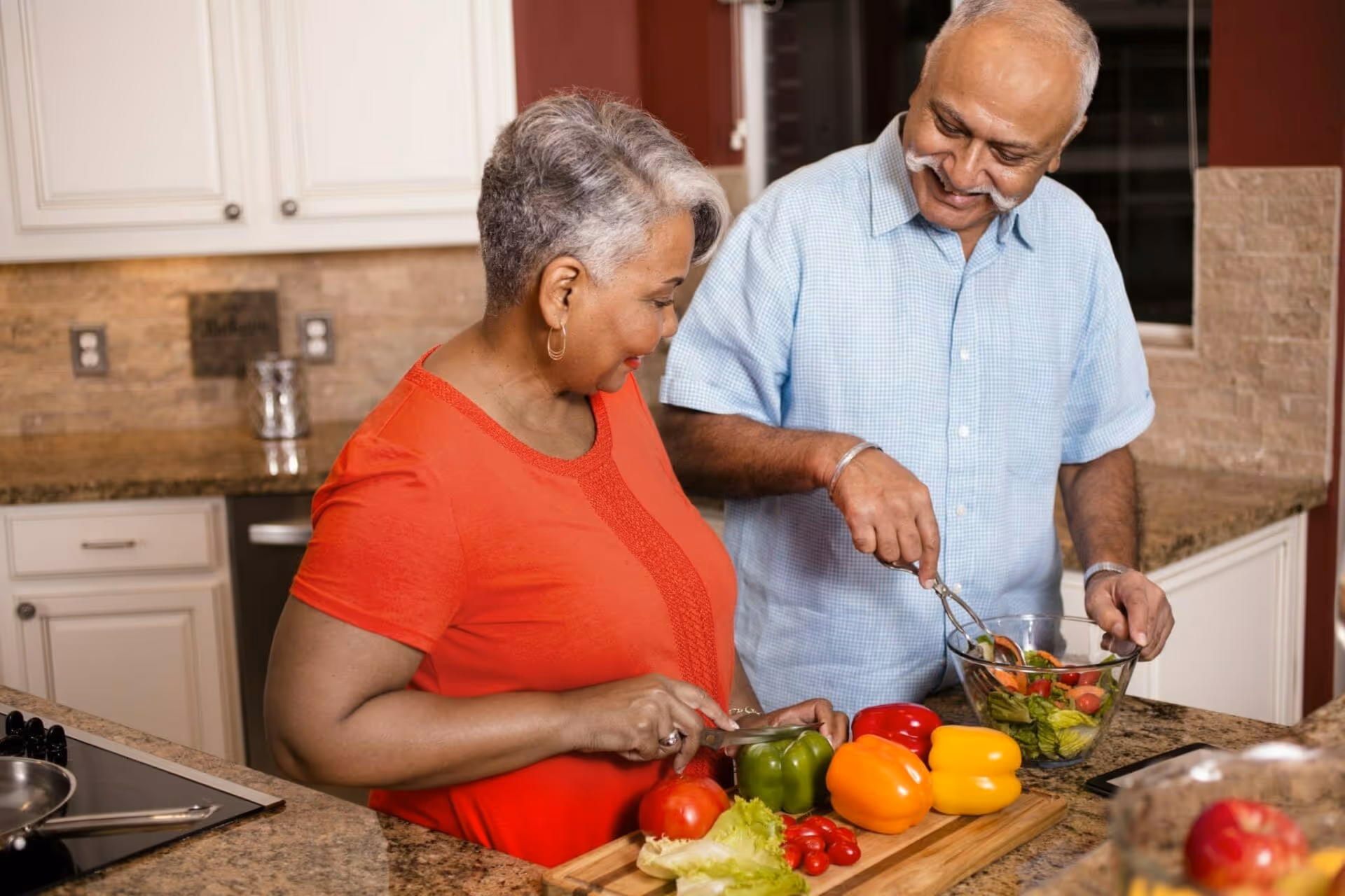 Two older adults prepare a salad together in a kitchen with colorful bell peppers and other vegetables on the counter.