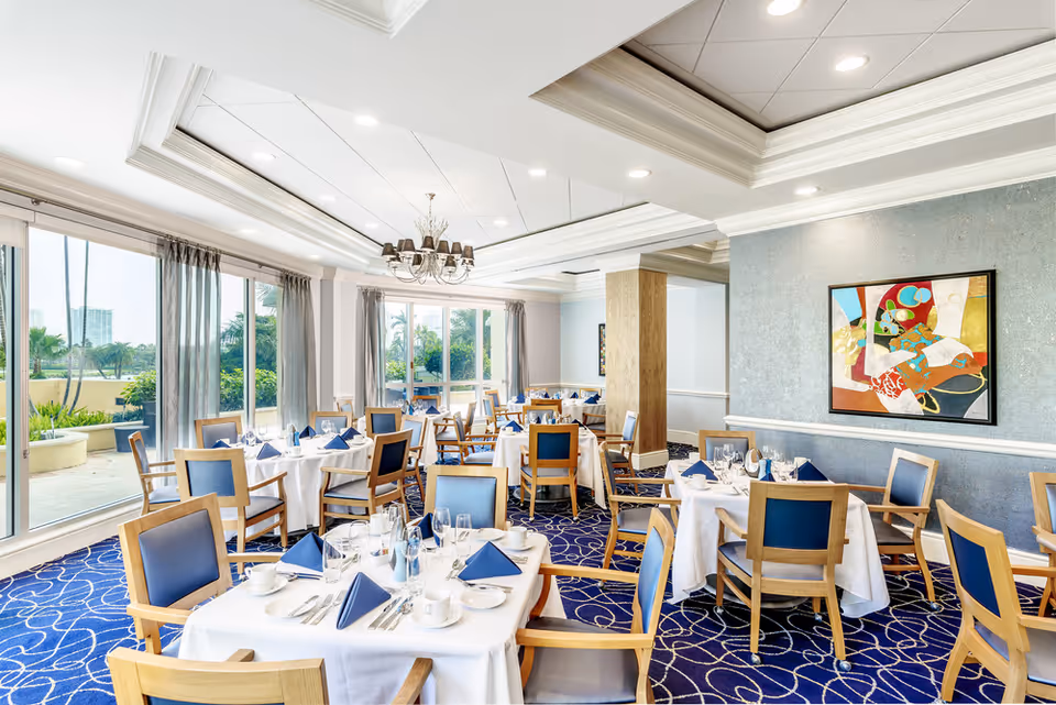 Bright and elegant dining room with multiple tables set with white tablecloths, blue napkins, glassware, and silverware. The room features large windows with sheer curtains allowing natural light to fill the space, blue patterned carpet, wooden chairs with blue cushions, a chandelier, and a colorful abstract painting on the wall.