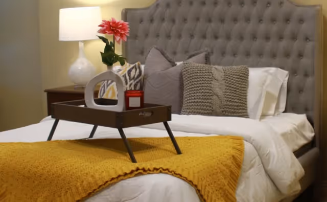 A styled bedroom with a tufted headboard, decorative pillows, and a tray holding a flower and candle on a bed with a mustard throw.