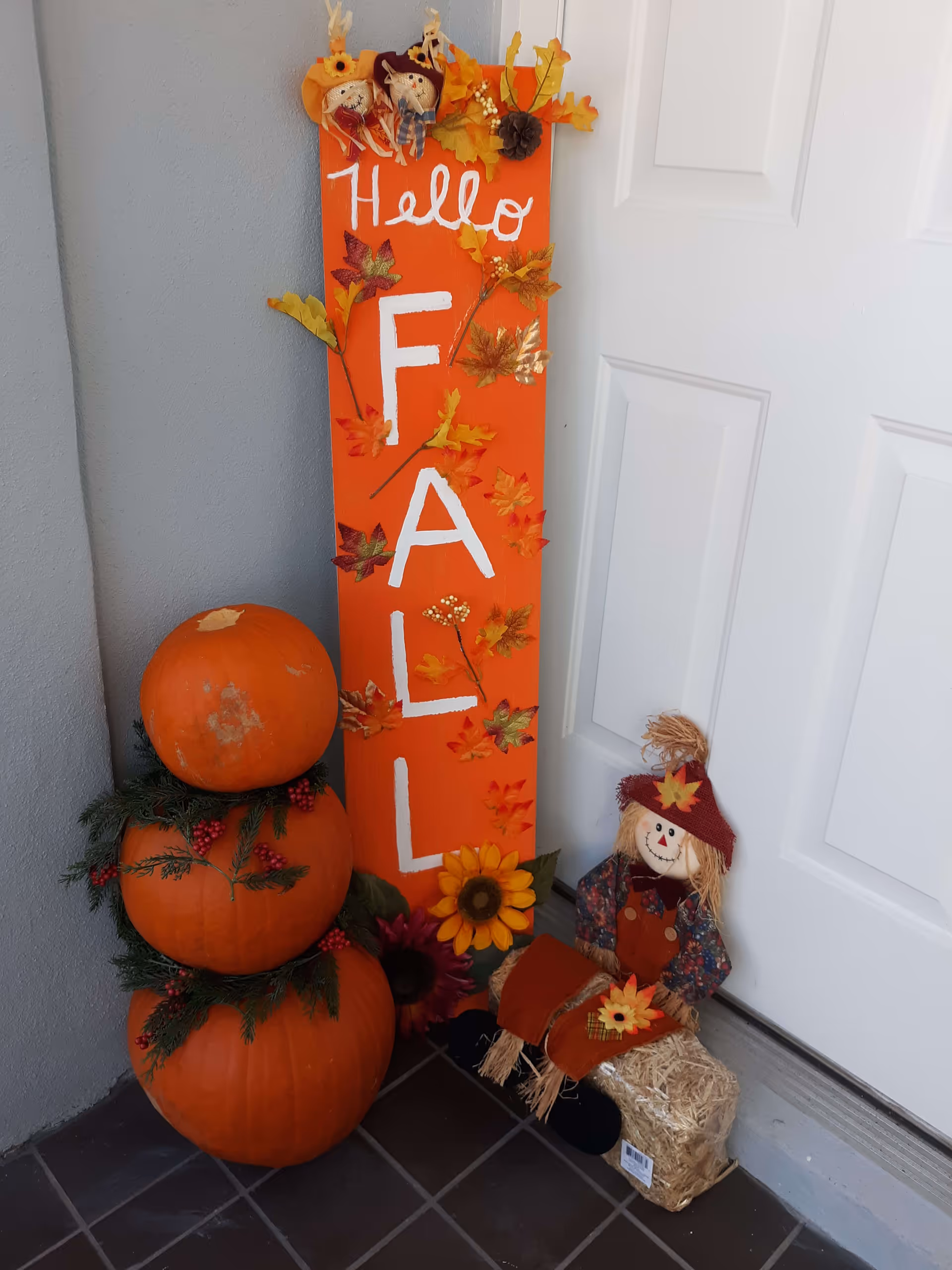 A fall-themed decoration arrangement by a white door, featuring a vertical orange sign with the words 'Hello FALL' written in white and adorned with artificial autumn leaves and small scarecrow figures. Next to the sign is a stack of three pumpkins decorated with green foliage and red berries. A small scarecrow doll dressed in autumn colors sits on a bale of straw beside the sign.