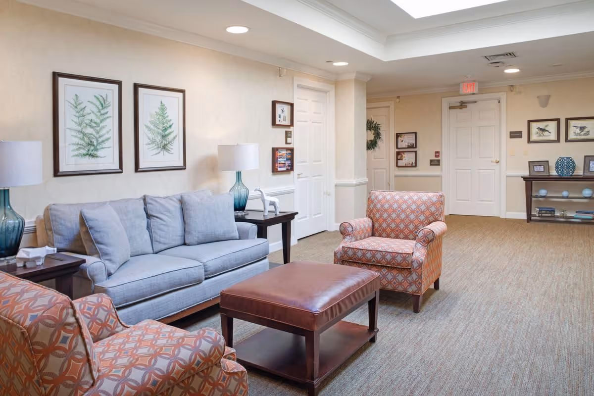 Well-lit common room with a light-gray sofa, patterned armchairs, a leather ottoman coffee table, side tables with lamps, framed botanical prints, and a console table against the wall.