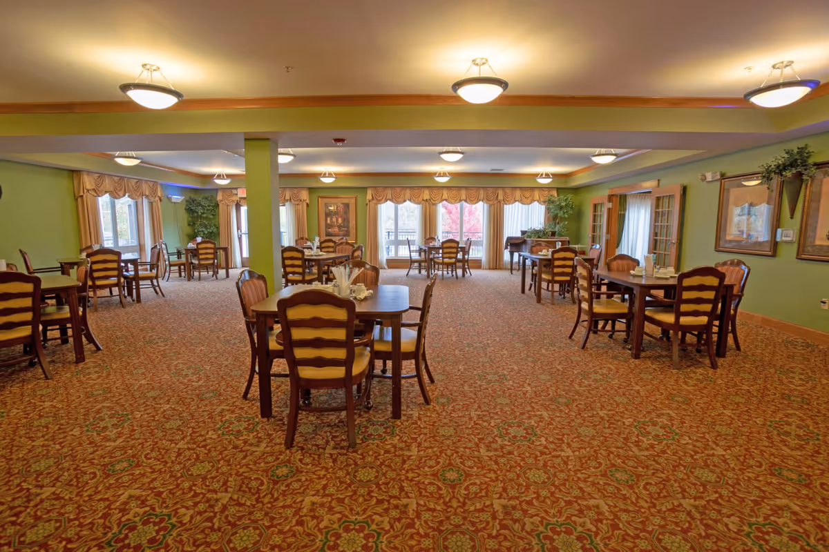 A spacious dining room with multiple wooden tables and chairs arranged neatly on a patterned carpet. The walls are painted green with framed artwork and plants for decoration. Large windows with curtains allow natural light to brighten the room.