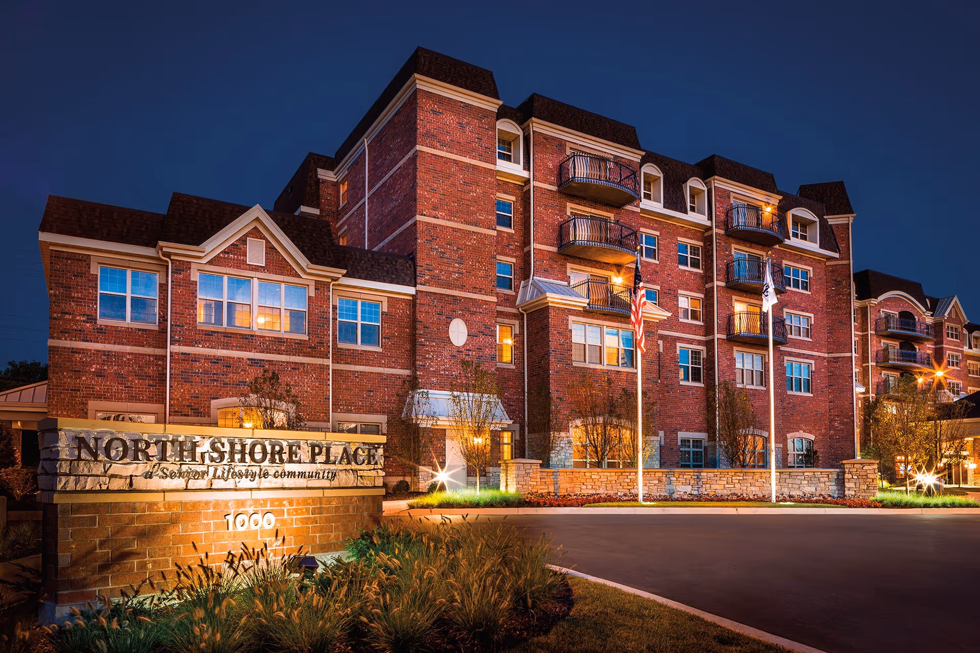 Exterior view of North Shore Place, a senior living community building, illuminated at dusk with multiple windows, balconies, and flags in front.