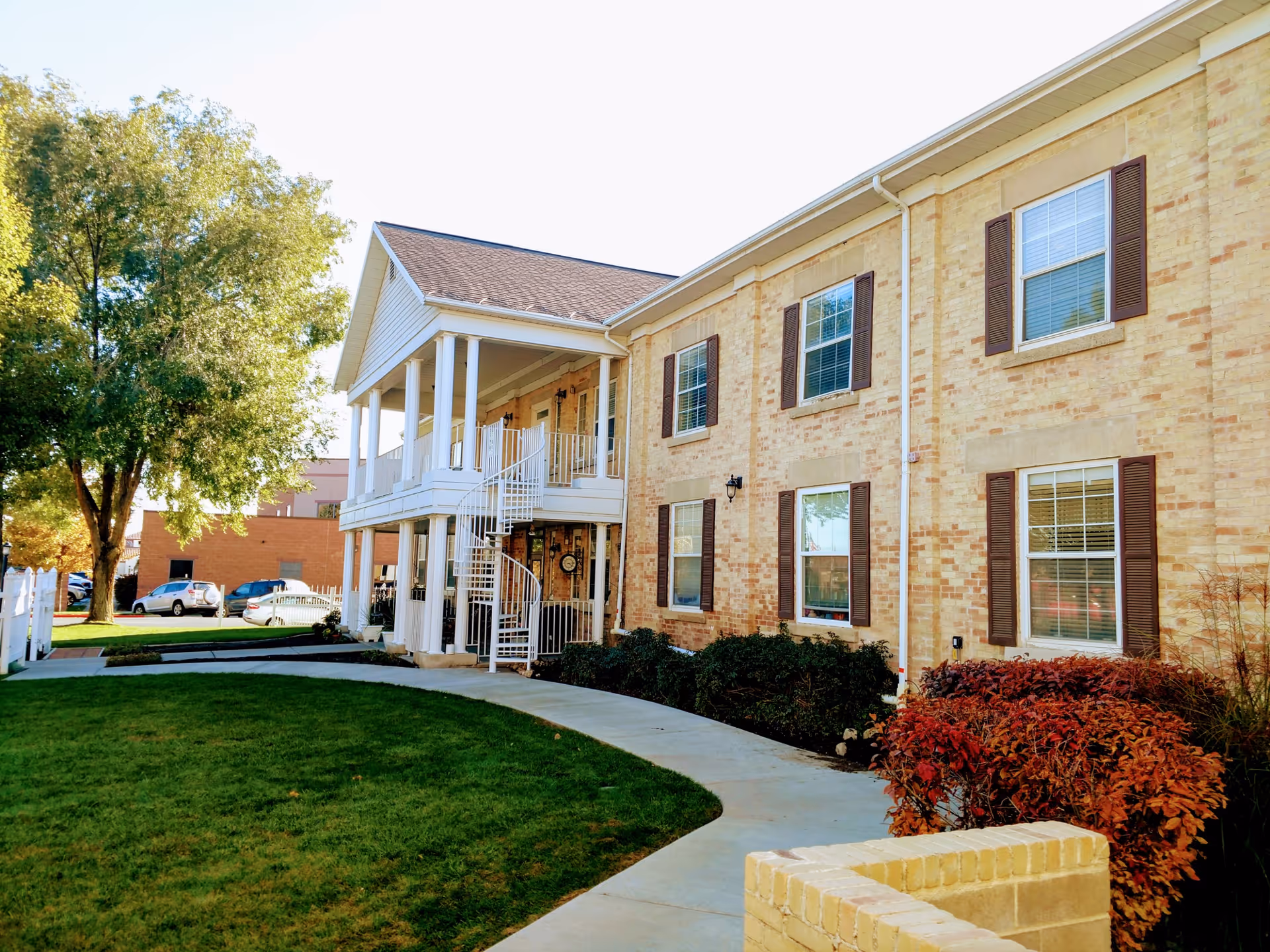 Two-story brick senior living building with a covered porch, exterior spiral staircase, walkway, and landscaped lawn.