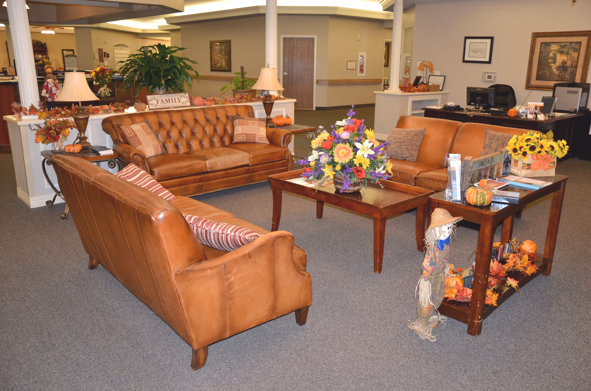 A cozy seating area in a nursing and rehabilitation facility featuring three brown leather sofas arranged around a wooden coffee table with a colorful floral arrangement. There is a side table with brochures, a small scarecrow decoration, and autumn-themed decor including pumpkins and flowers. The background shows a reception desk with office equipment and framed artwork on the walls.