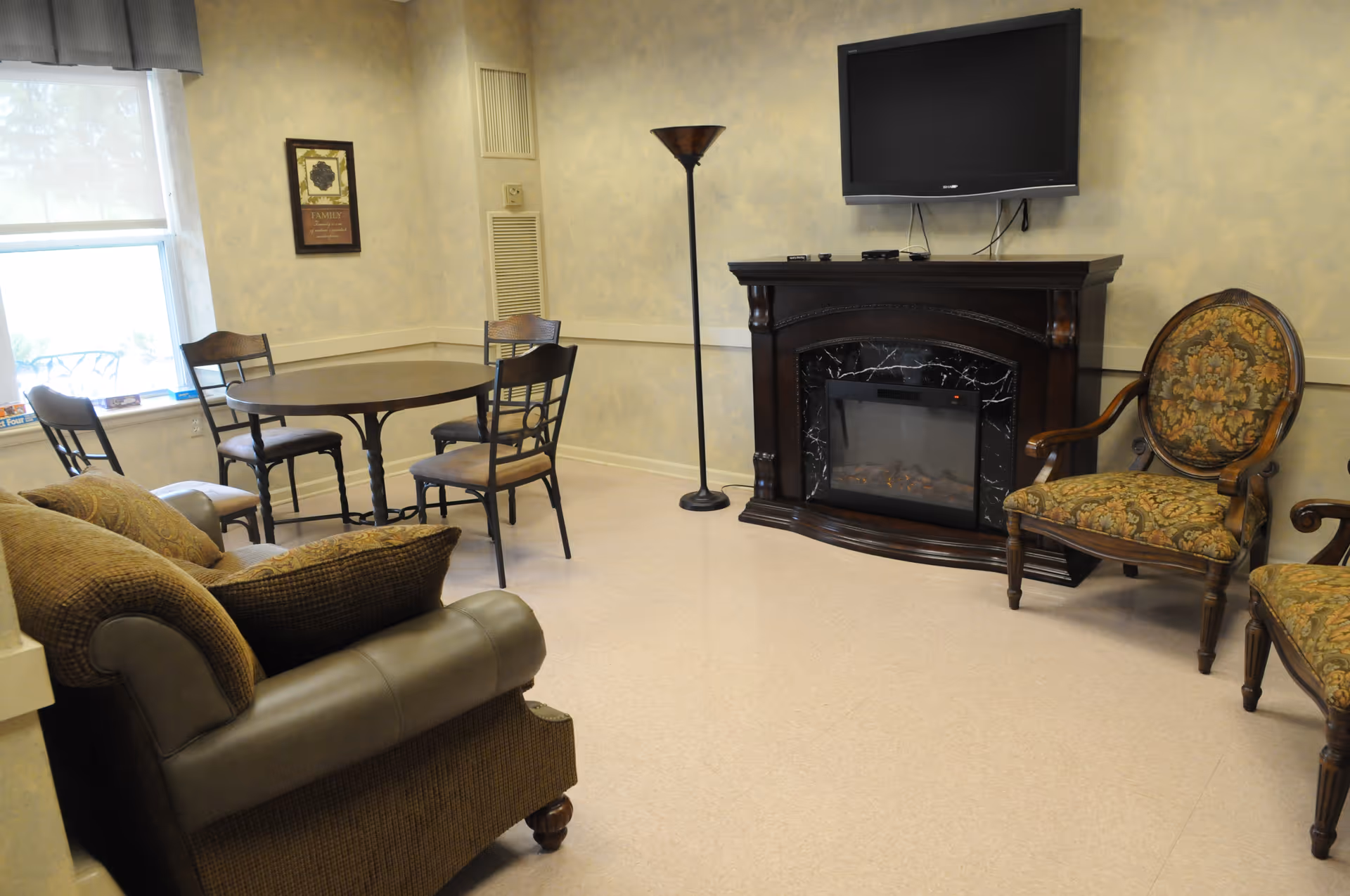 A cozy living room area with a brown cushioned armchair, a round wooden table with four chairs, a tall floor lamp, a dark wooden electric fireplace with a mounted flat-screen TV above it, and two upholstered armchairs with floral patterns. A window with a valance lets in natural light.