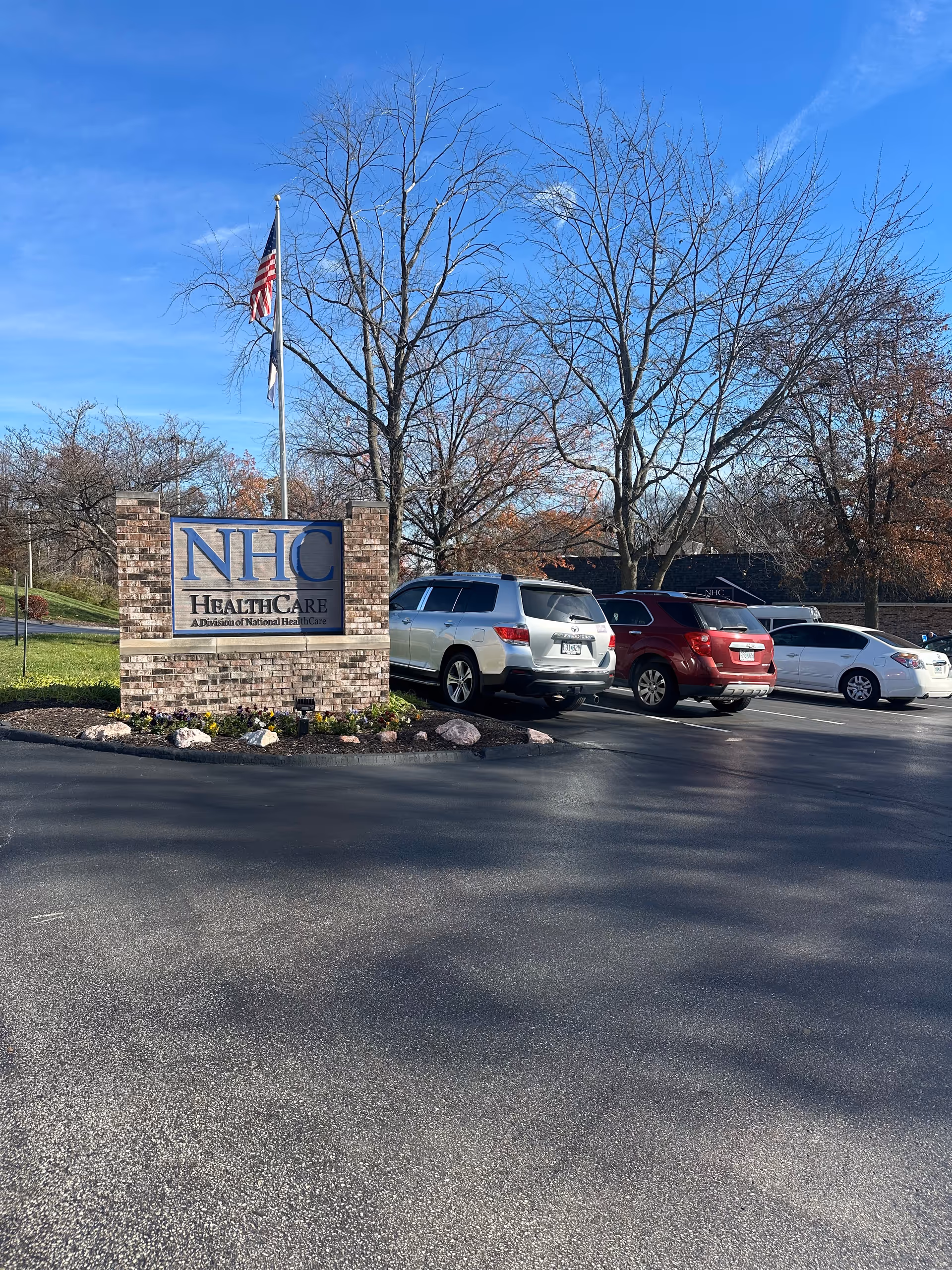 Brick entrance sign reading "NHC HealthCare" beside a parking lot with several cars, a flagpole, and leafless trees under a blue sky.