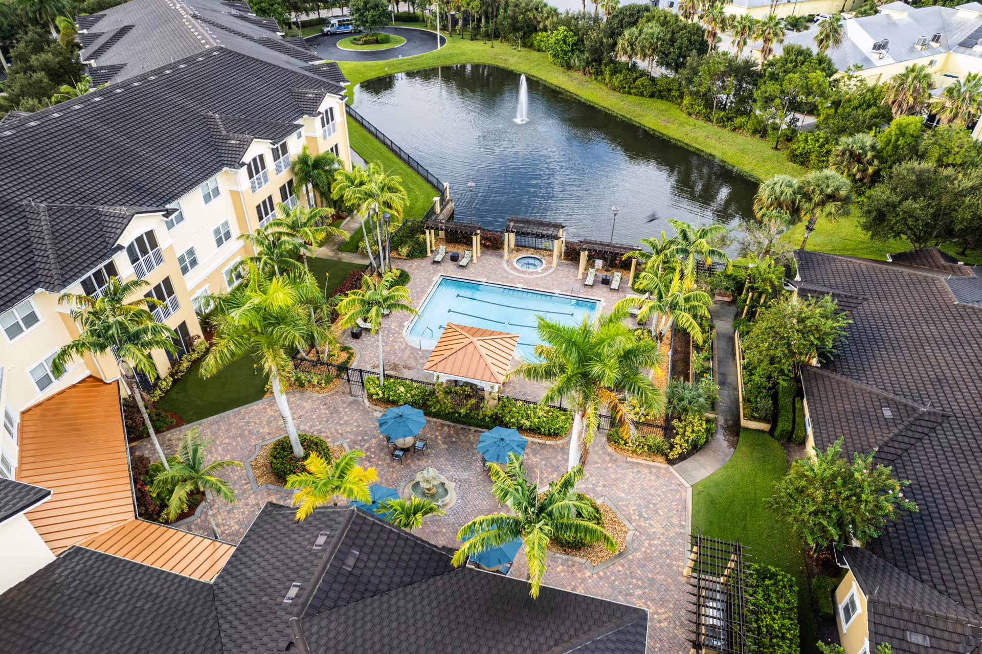 Aerial view of a courtyard with a swimming pool, hot tub, patio seating and palm trees beside a pond with a fountain, surrounded by apartment buildings.