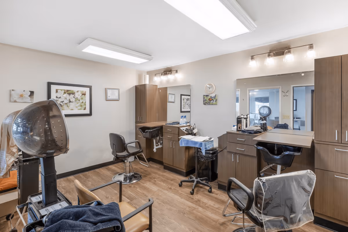 Interior view of a hair salon area within a senior living facility, featuring salon chairs, hair drying equipment, large mirrors, wooden cabinets, and framed floral artwork on the walls.