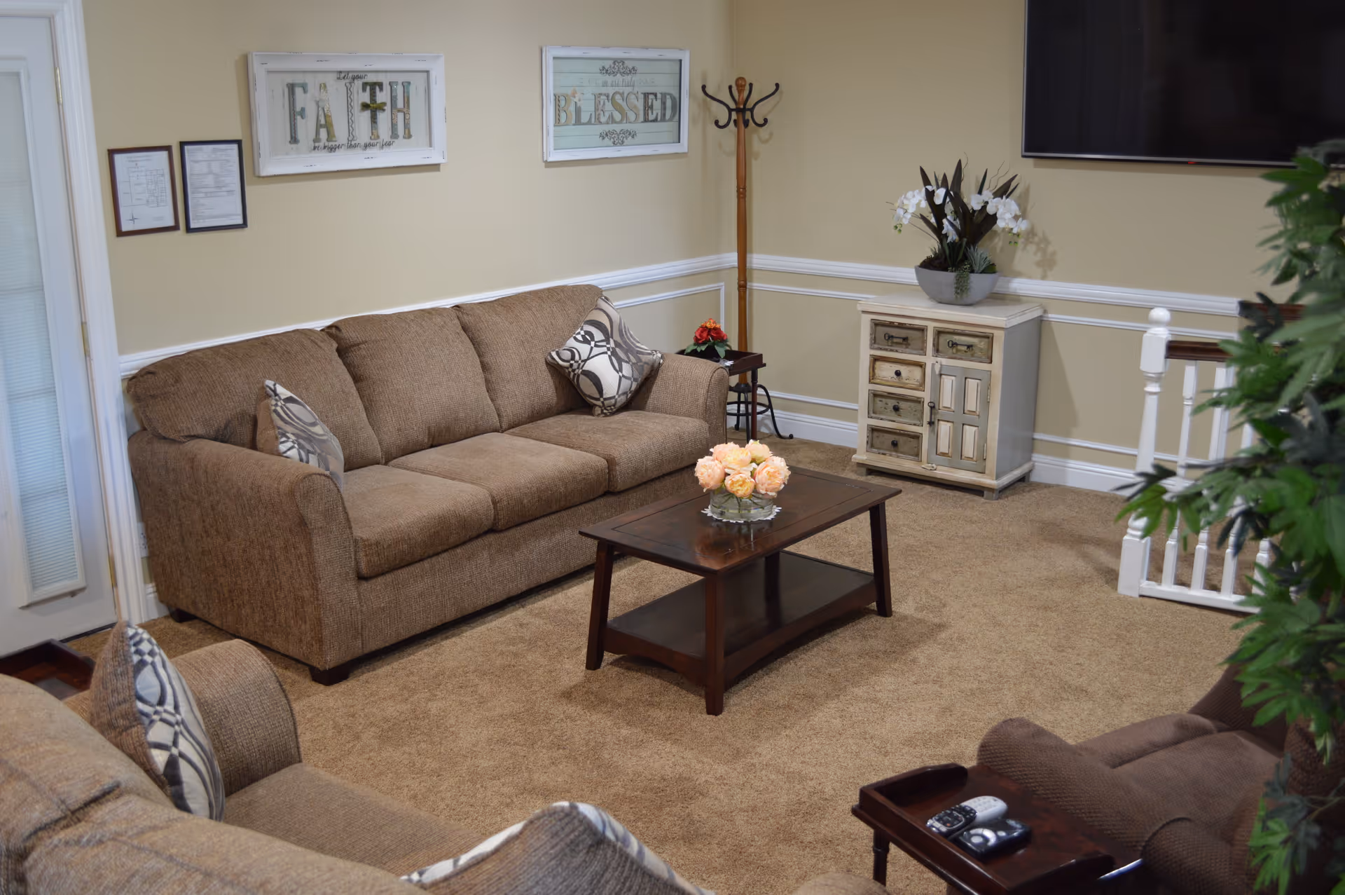 A cozy living room with brown sofas, a wooden coffee table topped with flowers, wall art reading 'FAITH' and 'BLESSED', and a small cabinet beneath a wall-mounted TV.