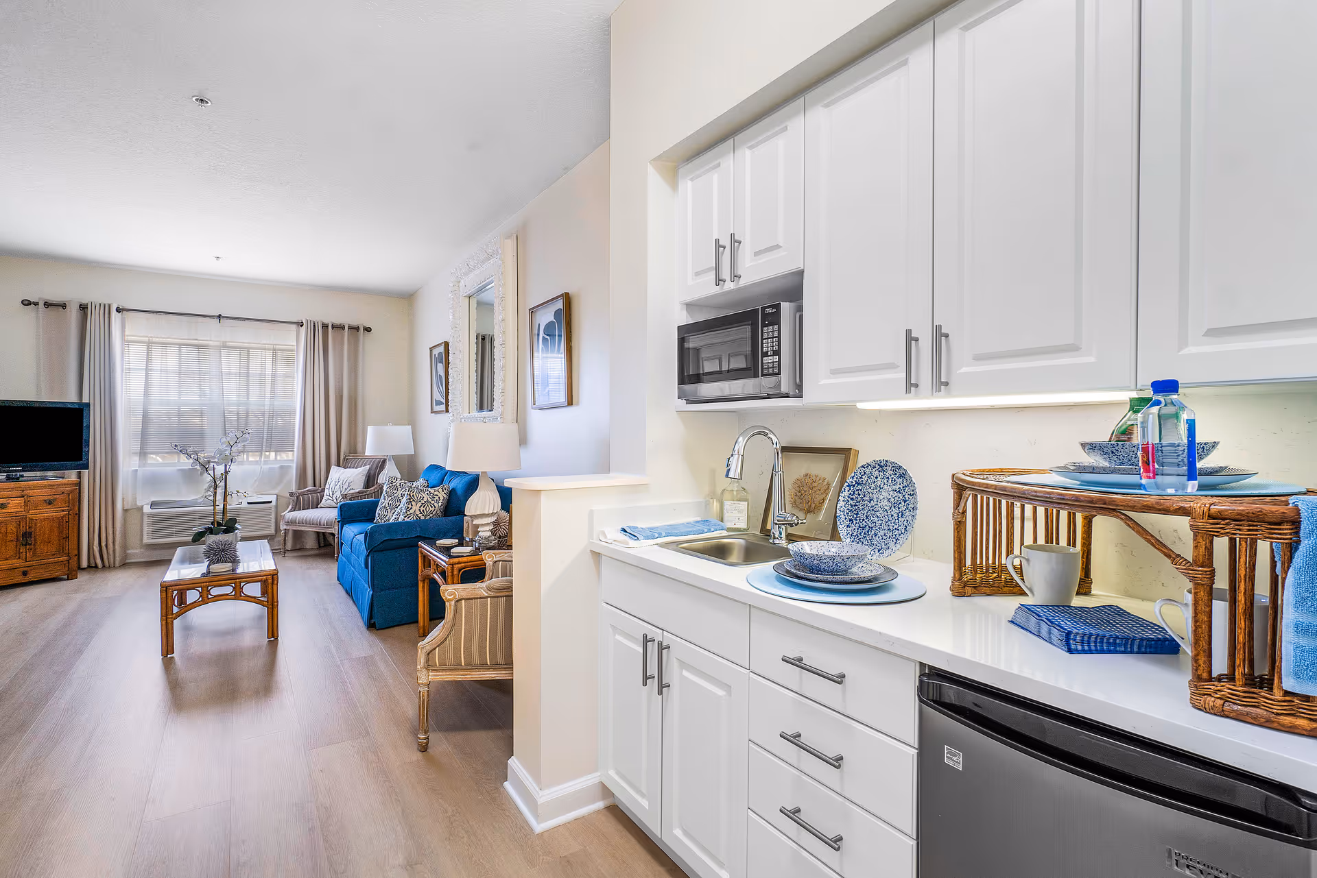 Bright open living area with a kitchenette, white cabinets, a blue sofa, coffee table and TV.