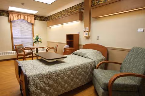 A furnished senior living bedroom with a bed, chair, and small table by a window