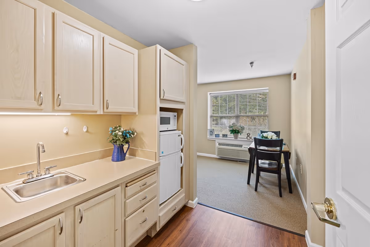 A small kitchen area with light wood cabinets, a stainless steel sink, a microwave, and a mini refrigerator. A blue vase with flowers is placed on the countertop. Beyond the kitchen is a dining area with a table and two chairs near a window with blinds, decorated with plants and framed photos.