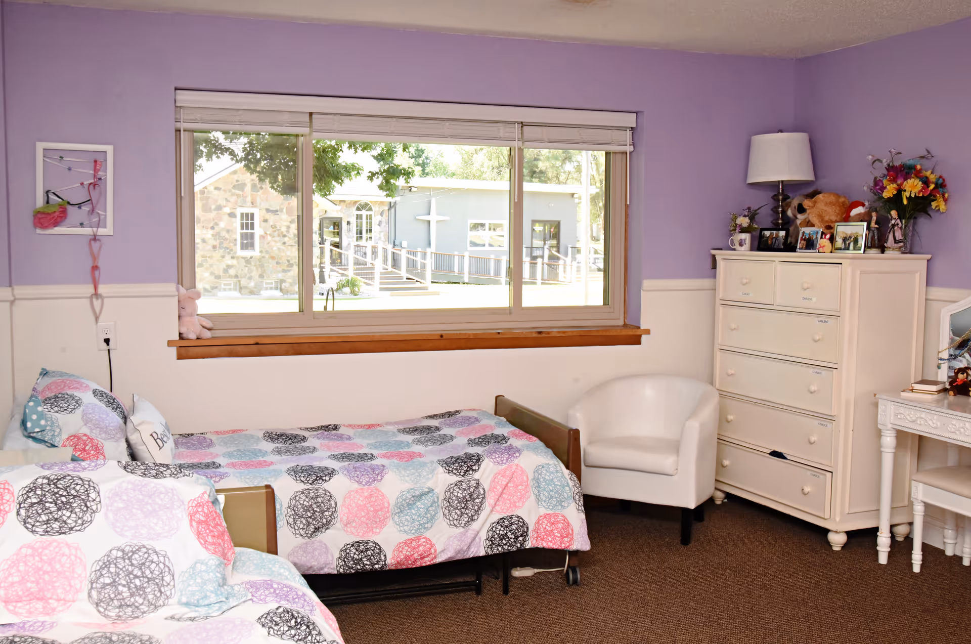 A cozy bedroom with two twin beds covered in colorful circular patterned bedspreads. The walls are painted lavender on the upper half and white on the lower half. A large window shows an outdoor view of a stone building with a white cross. There is a white armchair, a white dresser with a lamp, framed photos, flowers, and a teddy bear on top. A small white desk with a chair is partially visible on the right side.