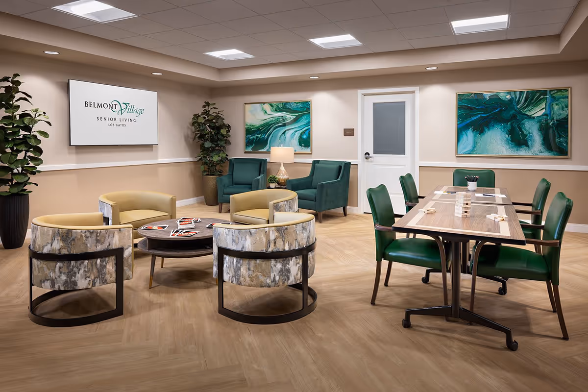 Well-lit senior living common room with circular lounge chairs, green armchairs, a games table, wall artwork, and a Belmont Village sign.