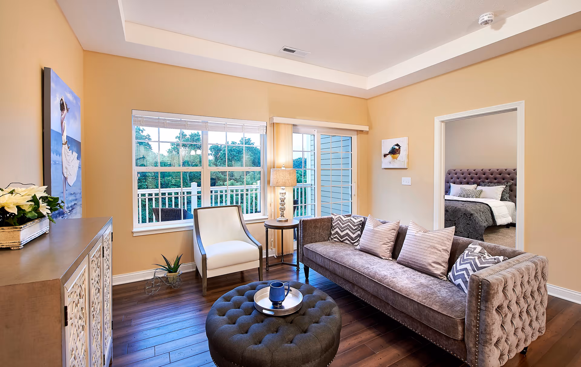 A cozy living room with a tufted gray sofa adorned with patterned and solid pillows, a round tufted ottoman with a tray and cup, a white armchair, a side table with a lamp, and large windows showing greenery outside. A doorway leads to a bedroom with a tufted headboard and neatly made bed.