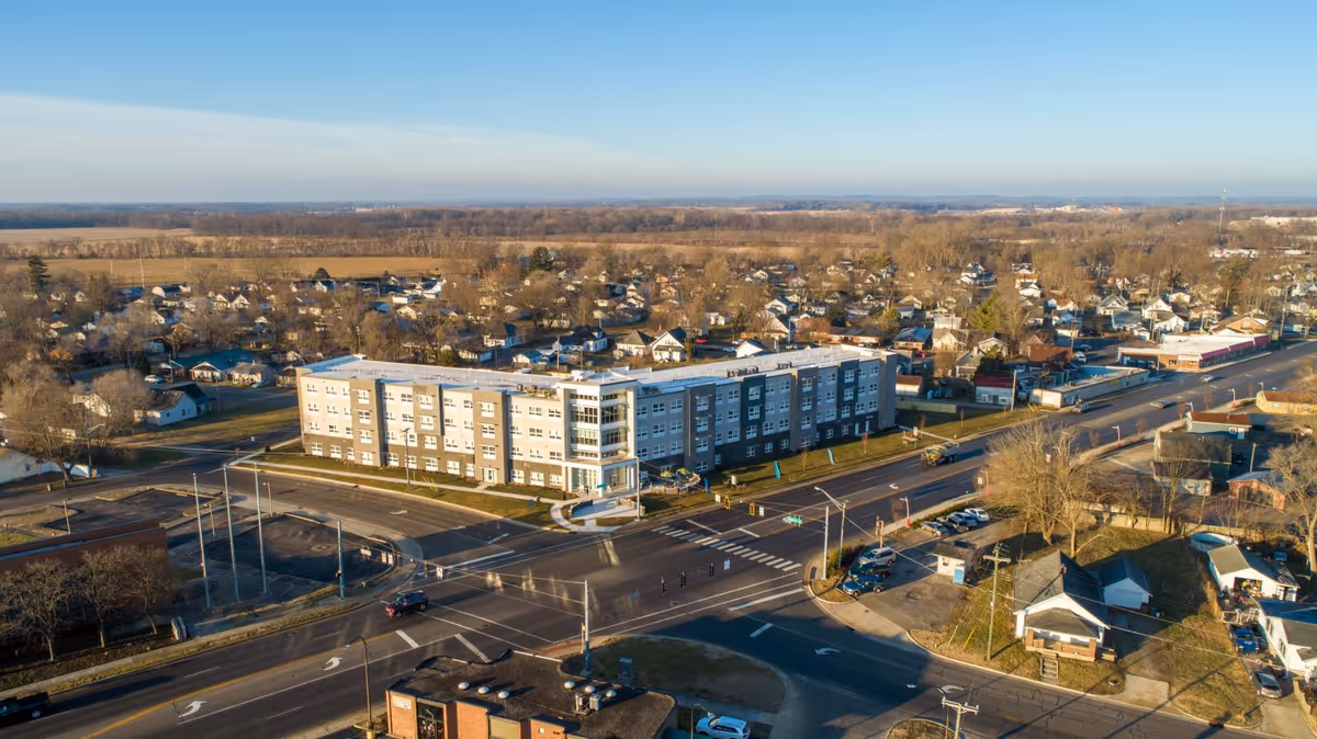Aerial view of a large, modern four-story senior living facility named Vivera Senior Living of Columbus, situated at a busy intersection with surrounding residential houses and open land in the background under a clear sky.