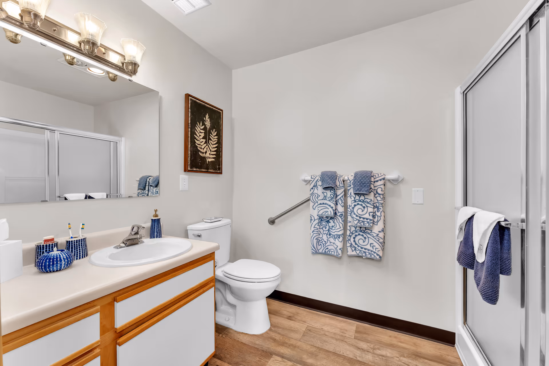 A clean and well-lit bathroom with a white sink and countertop, wooden cabinetry, a large mirror with three light fixtures above it, a toilet, a framed botanical artwork on the wall, two patterned blue and white towels hanging on a towel rack, and a glass-enclosed shower with two dark blue towels hanging on the door handle. The floor is wood-style, and the walls are painted white.