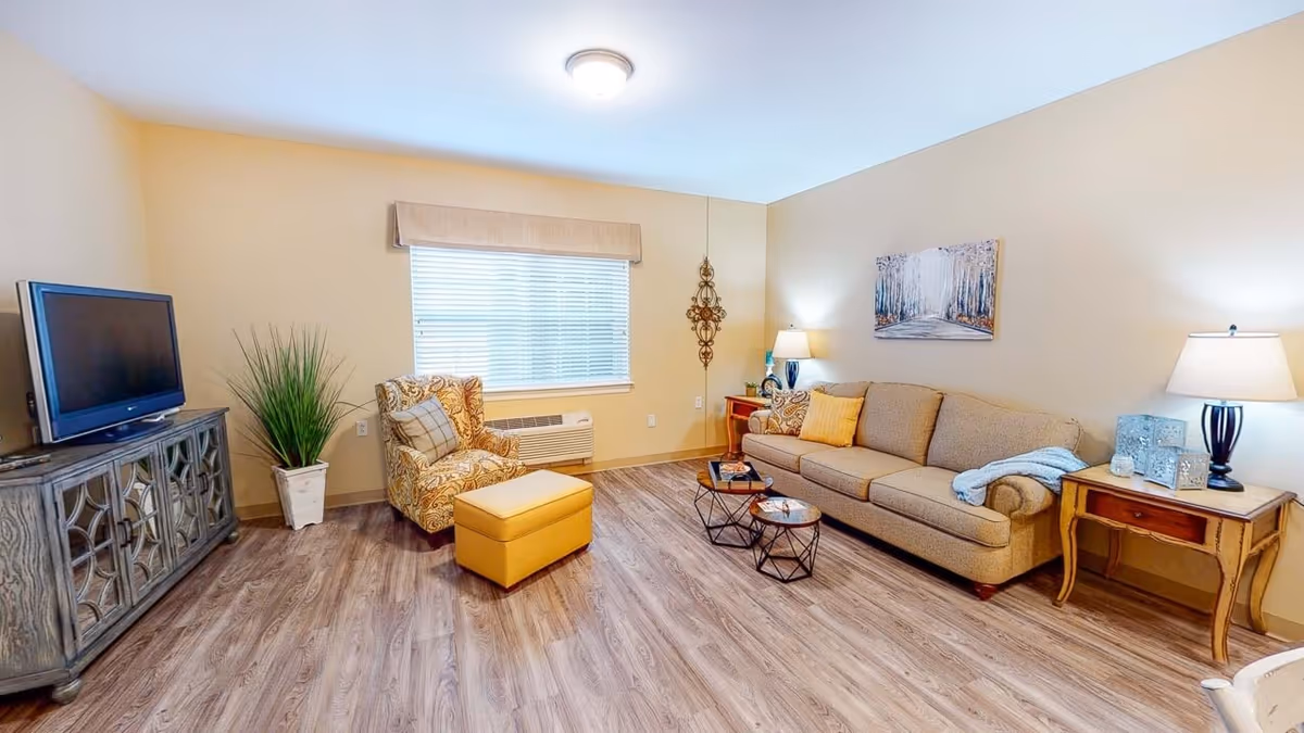 A cozy living room with a beige sofa, a patterned armchair with a matching ottoman, two small round coffee tables, a wooden side table with a lamp, a TV on a decorative cabinet, a potted plant, and a window with blinds and a valance. The walls are light beige and the floor is wood.