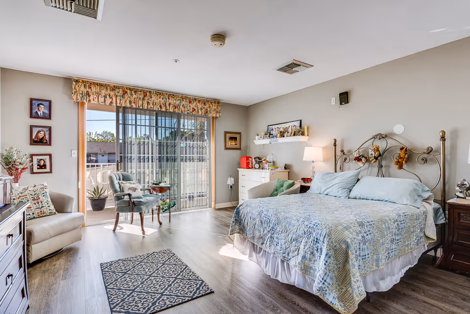 A bright and cozy bedroom with a large bed covered in a blue and beige patchwork quilt. The room has wooden flooring, a patterned rug, and a sliding glass door leading to a balcony with outdoor furniture. There are two armchairs, a small table, a dresser with various items on top, and framed photos on the walls. The room is decorated with stuffed animals hanging on the bed frame and has a warm, inviting atmosphere.