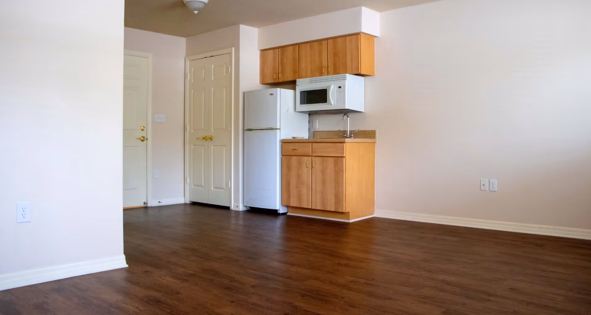 Empty studio apartment interior with wood floors and a small kitchenette featuring a refrigerator, microwave, sink, and cabinets.
