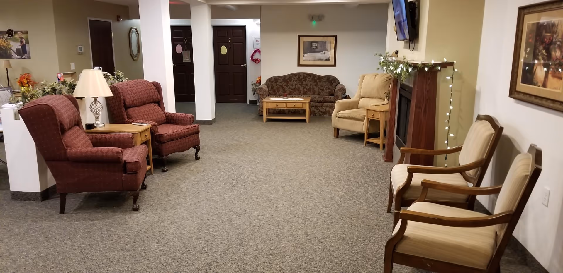 Carpeted communal sitting area with upholstered armchairs, a sofa, a fireplace with string lights, a wall-mounted TV, and a hallway with numbered doors.