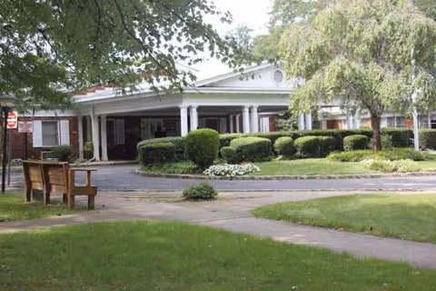 White, single-story building with a covered entrance, surrounded by hedges and trees.