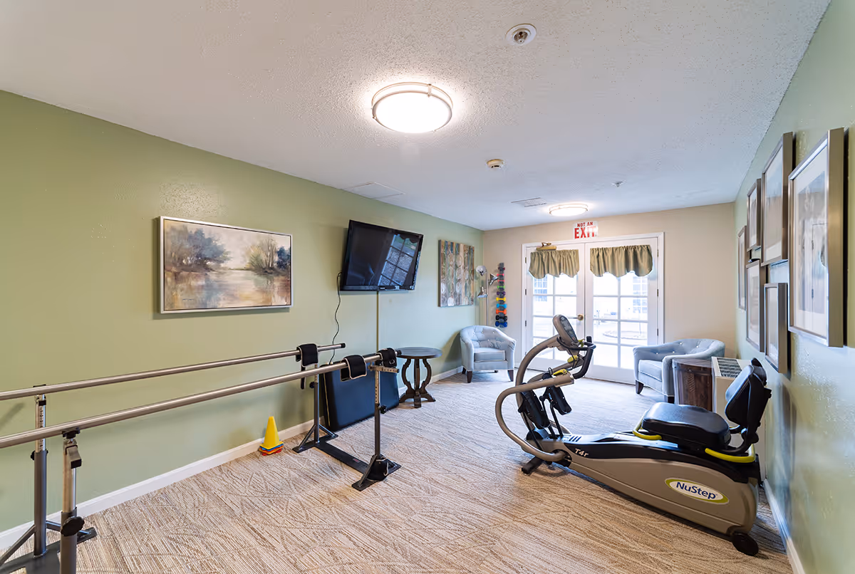 A small exercise room with green walls and carpeted floor featuring parallel bars for walking support, a NuStep exercise machine, two armchairs, a small round table, a wall-mounted TV, and framed artwork. Double glass doors with green curtains lead outside, and there is a rack with colorful dumbbells near the back wall.