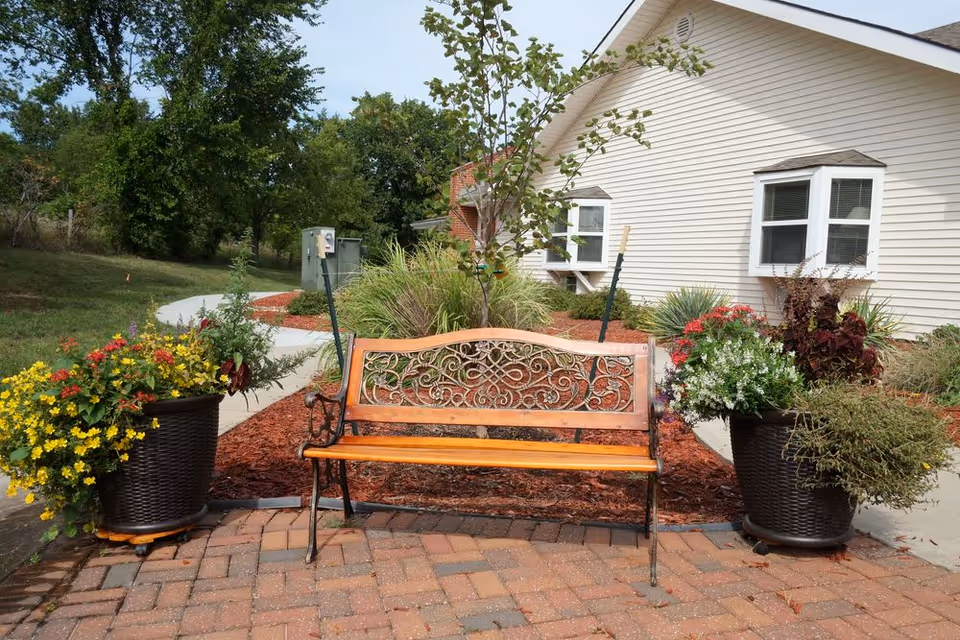 A decorative wooden bench with ornate metal backrest sits on a brick-paved area between two large planters filled with colorful flowers and greenery. Behind the bench is a landscaped garden with mulch, shrubs, and a small tree, adjacent to a light-colored building with white-framed windows. A concrete pathway curves through the garden area, and trees and grass are visible in the background under a partly cloudy sky.