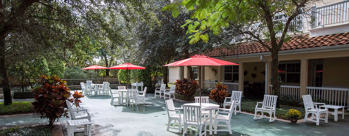 Outdoor patio area with several white tables and chairs, some shaded by red umbrellas. The patio is surrounded by trees and plants, with a building featuring a tiled roof and windows in the background.