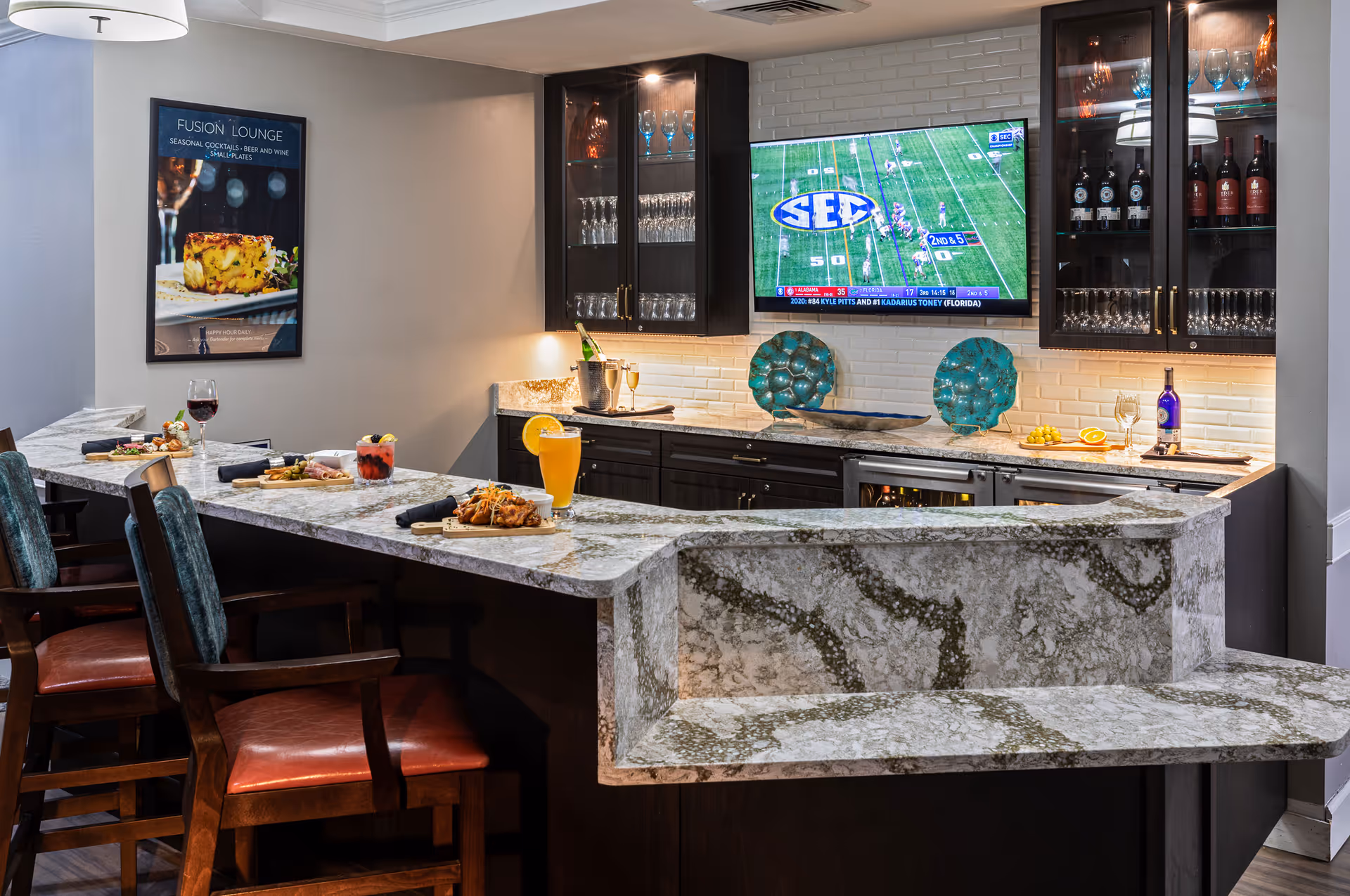 Interior view of a lounge bar area with a marble countertop and several high chairs. The countertop has various drinks and small plates of food. Behind the bar are dark wooden cabinets with glass doors displaying wine glasses and bottles. A flat-screen TV mounted on a white tiled wall shows a football game. A framed poster on the wall reads 'Fusion Lounge Seasonal Cocktails, Beer and Wine, Small Plates.'