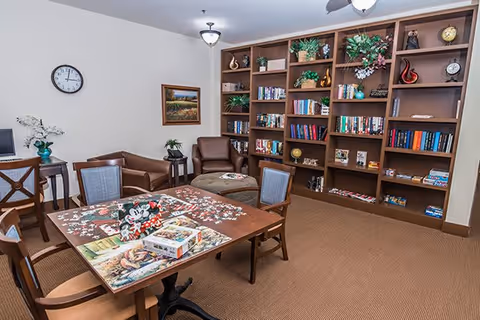 A cozy common room with a large wooden bookshelf filled with books and decorative items against the wall. In front of the bookshelf are two brown armchairs and a matching ottoman. A wooden table with four chairs surrounds a partially completed jigsaw puzzle featuring Disney characters. The room has beige carpet, a wall clock, a framed painting, and a small side table with a plant and flowers.