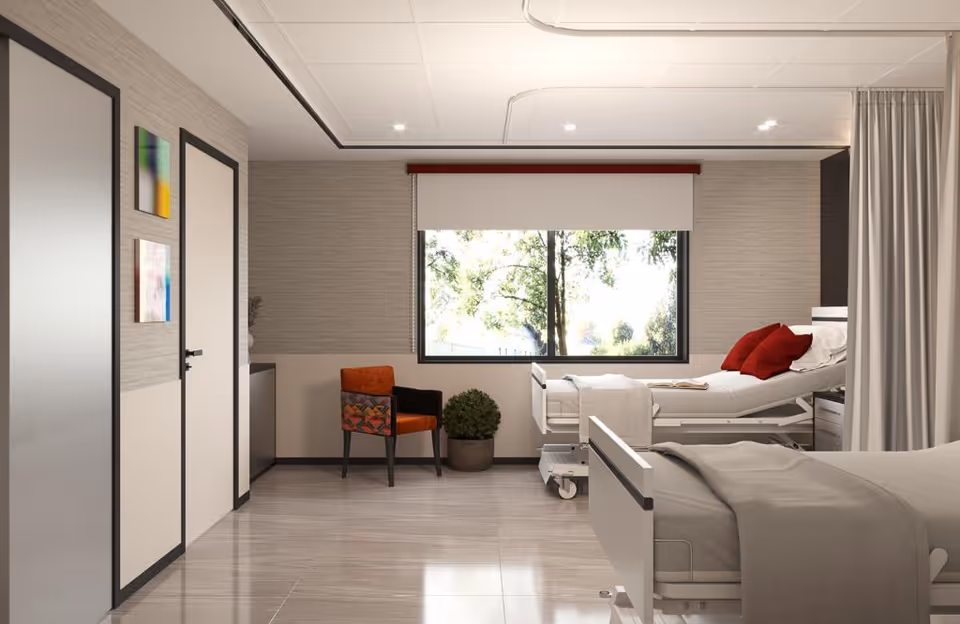 A bright and clean nursing home room with two hospital beds, one near a large window with red pillows and a beige blanket, and the other partially visible in the foreground. There is an orange chair with a patterned backrest and a potted plant near the window. The room has light-colored walls, tiled floors, and a ceiling with recessed lighting.