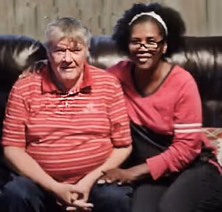 An elderly man wearing a red striped polo shirt sitting on a dark leather couch next to a woman with glasses and a white headband, wearing a red and black top. They are indoors and sitting close together, smiling at the camera.