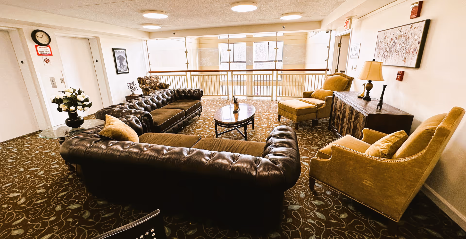 Cozy communal seating area with tufted leather sofas, upholstered armchairs, a coffee table, and large windows overlooking an atrium.