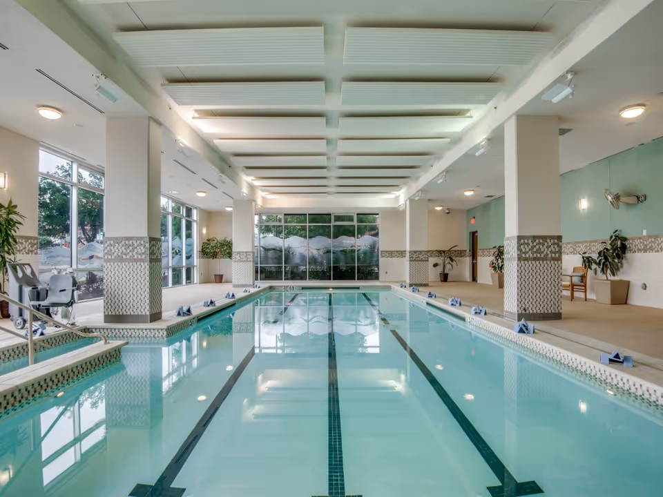 Indoor swimming pool area with clear water, tiled edges, and handrails. The room has large windows on one side letting in natural light, potted plants along the walls, and a ceiling with white acoustic panels. There are chairs and a pool lift for accessibility.